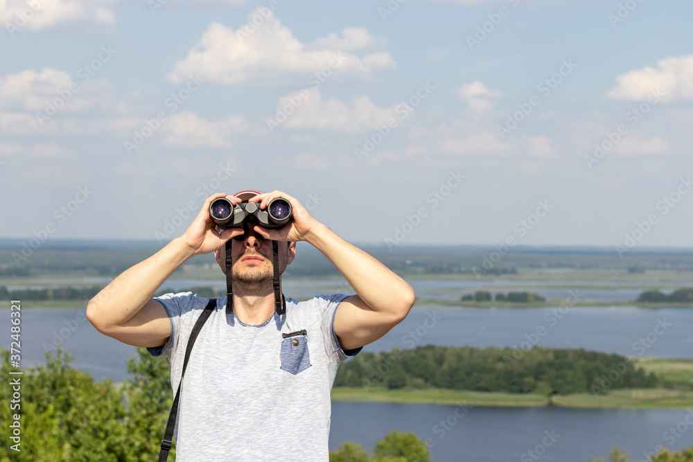 A man looks through binoculars at sky against a scenic view of a wide river on a hill on a sunny day A man looks through binoculars at sky against a scenic view of a wide river on a hill on a sunny day