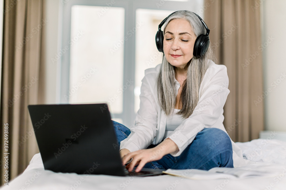 Horizontal shot of smiling gray haired senior woman, sitting on bed in home bedroom, typing on laptop, listening to music or video in headphones and working online or chatting with friend Horizontal shot of smiling gray haired senior woman, sitting on bed in home bedroom, typing on laptop, listening to music or video in headphones and working online or chatting with friend