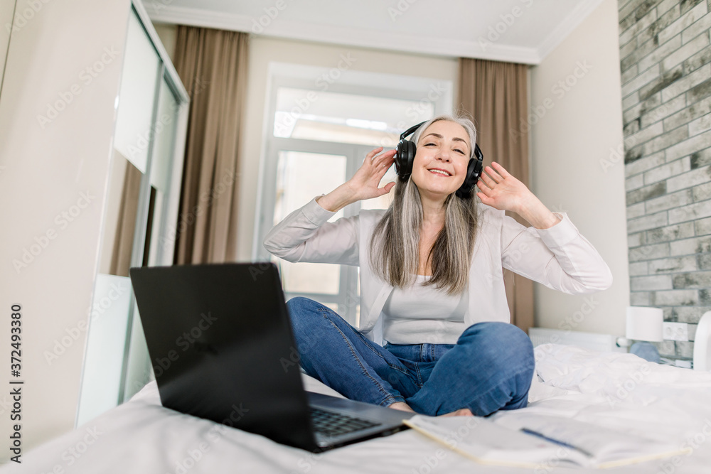 Modern elderly people concept. Happy senior lady with long gray hair having fun, enjoying listening to music in headphones, while sitting on the bed and searching information on laptop Modern elderly people concept. Happy senior lady with long gray hair having fun, enjoying listening to music in headphones, while sitting on the bed and searching information on laptop
