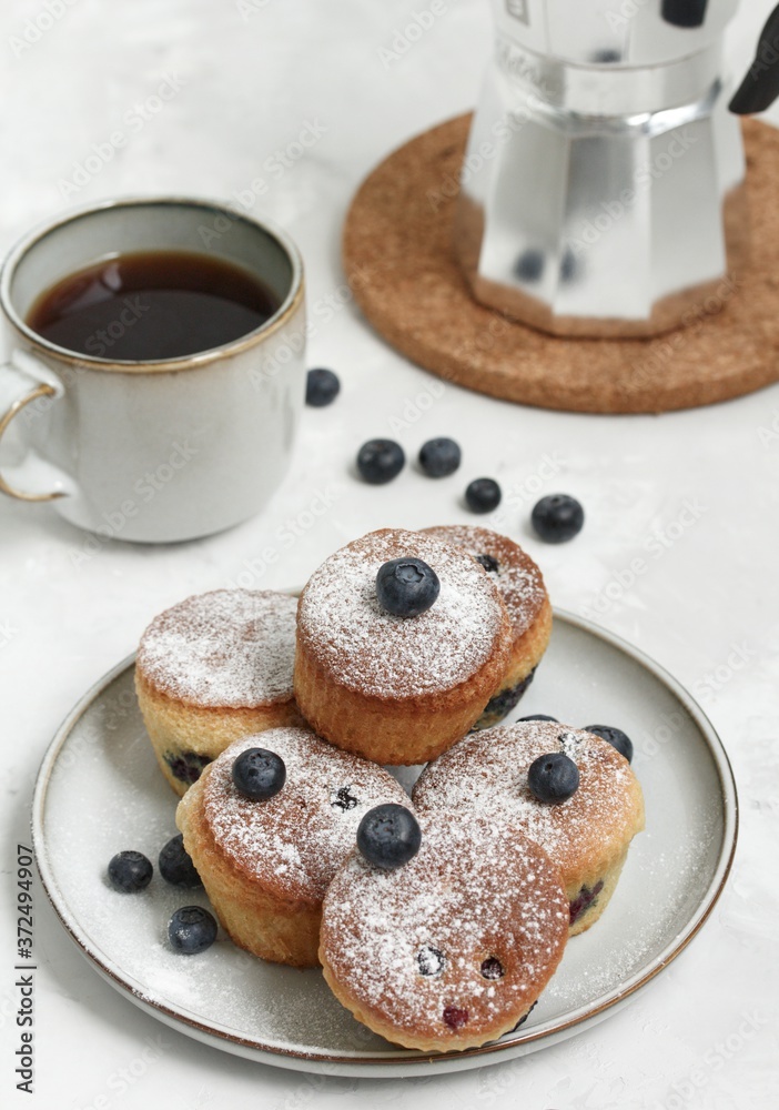 Muffins on a gray plate on a light table with a cup of coffee, blueberries and a coffee pot