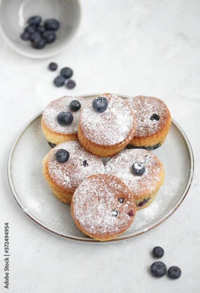 Muffins decorated with blueberries and powdered sugar on a light gray table. Blueberries are scattered on the table