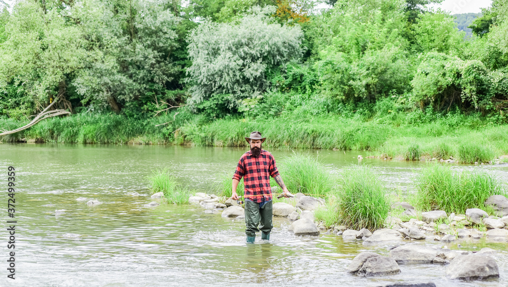 Satisfied fisher. Fishing requires you to be mindful and fully present in moment. Brutal man wear rubber boots stand in river water. Fisher masculine hobby. Fisher fishing equipment. Fish on hook