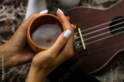 Cozy photo of woman's hands holding cup of tea with ukulele on backdrop. Fall or winter time concept.