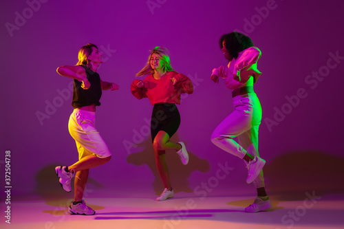 Fototapeta Naklejka Na Ścianę i Meble -  Flexible. Sportive girls dancing hip-hop in stylish clothes on purple-pink background at dance hall in green neon light. Youth culture, movement, style and fashion, action. Fashionable portrait.