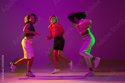 Flexible. Sportive girls dancing hip-hop in stylish clothes on purple-pink background at dance hall in green neon light. Youth culture, movement, style and fashion, action. Fashionable portrait.