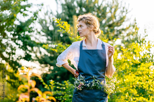 Beautiful portrait of smiling middle-aged woman with wild flowers in pocket, holding secateurs in garden. She is absolutely happy. Slow living, gardening hobby concept
