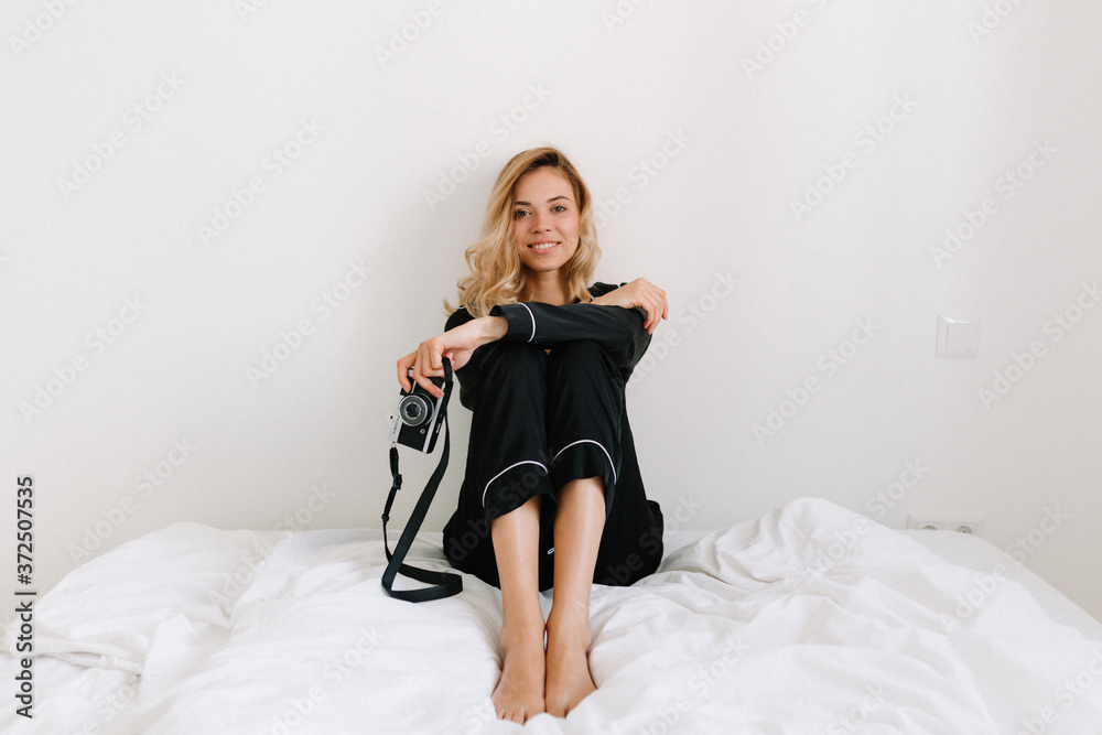 Naklejka premium Full-lenght photo of lovely tender girl with blonde hair wearing home suit sitting on the white bed with smile and posing at camera. Happy morning at home