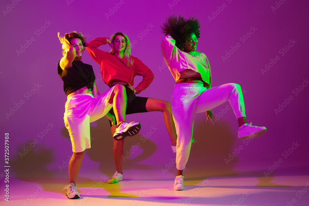 Flexible. Sportive girls dancing hip-hop in stylish clothes on purple-pink background at dance hall in green neon light. Youth culture, movement, style and fashion, action. Fashionable portrait.