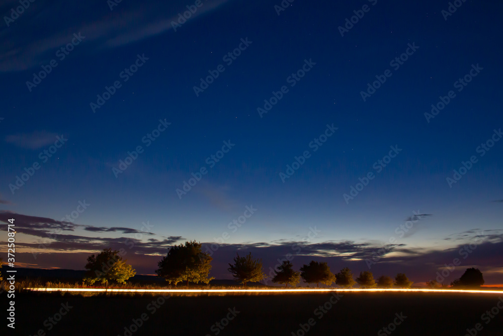 Headlights of a car driving on a country road and illuminating trees during sunset, long exposure