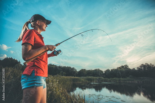 Fototapeta Cute caucasian woman is fishing with rod on the summer lake