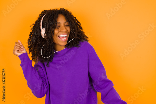 Young woman  wearing casual clothes against orange wall, dancing and listening music with headphones. © Jihan