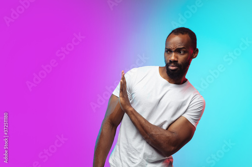 Avoiding. African-american young man's portrait on gradient studio background in neon. Beautiful male model in casual style, white shirt. Concept of human emotions, facial expression, sales, ad.