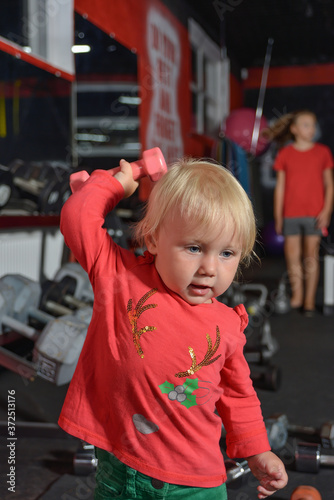 A small child in the gym raises a dumbbell up. Little girl goes in for sports.