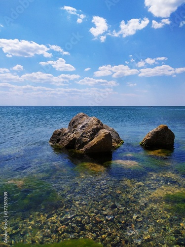 Beautiful seascape: pebble beach, clear water, stones overgrown with green algae on the shore, blue sky with clouds, calm.