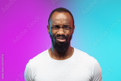 Close up offended. African-american young man s portrait on gradient background in neon. Beautiful male model in casual style  white shirt. Concept of human emotions  facial expression  sales  ad.