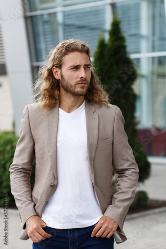 Business portrait of handsome curly smiling man with long hair in casual wear, standing on city street near business center. Confident businessman became successful.