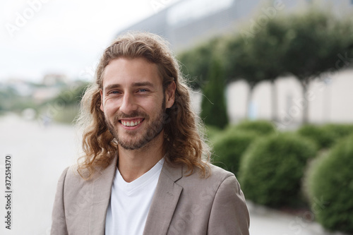 Business portrait of handsome curly smiling man with long hair in casual wear, standing on city street near business center. Confident businessman became successful.