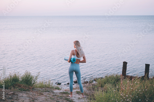 young slender woman in sportswear with a sports mat in her hands came to perform sports exercises on the beach © Mikhaylovskiy 
