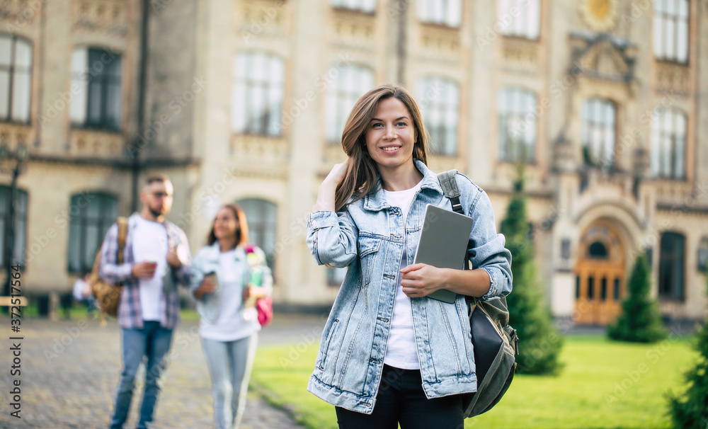Beautiful young student girl with a backpack and books is looking on camera on group of her friends background in university