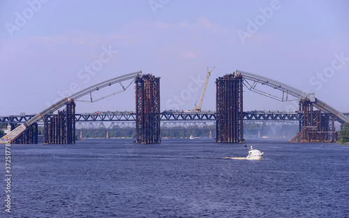 View of the unfinished bridge under constriction and pleasure boat floating on the river © Yurii Zushchyk