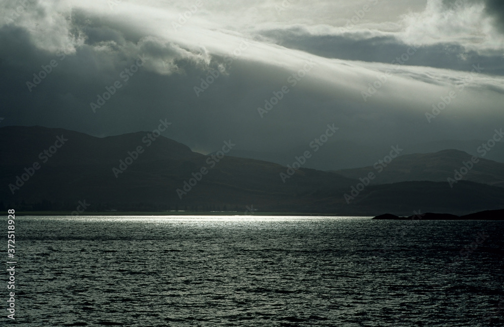 Sea of Mull in Scottish Highlands UK. View taken from Isle of Mull bank side. Stormy sky in late afternoon. White clouds with sunset reflecting on water. Mountains in background.