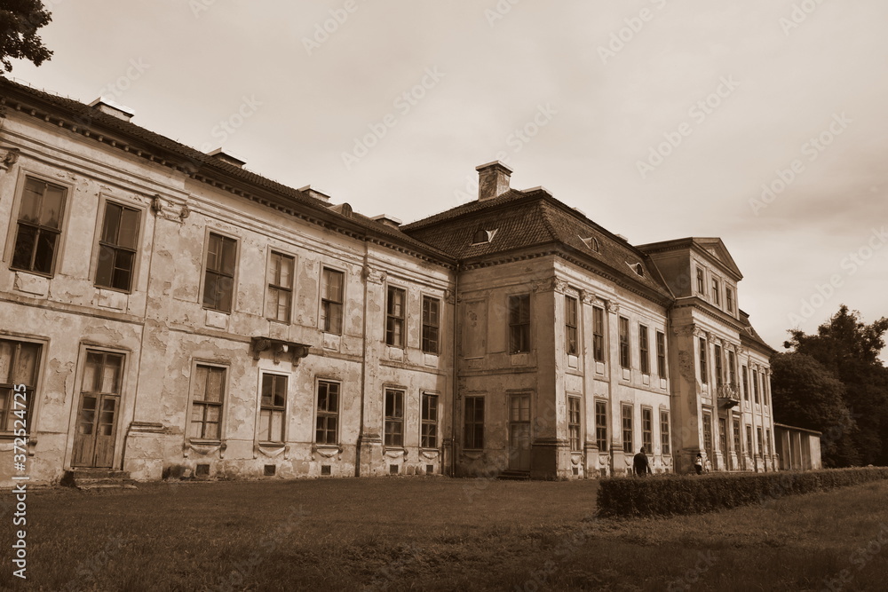 View of an old abandoned and slightly ruined mansion or palace with many windows, arch based entrance, and a slanted red roof surrounded with beautidul gardens, hedges, and tall trees seen in Poland View of an old abandoned and slightly ruined mansion or palace with many windows, arch based entrance, and a slanted red roof surrounded with beautidul gardens, hedges, and tall trees seen in Poland