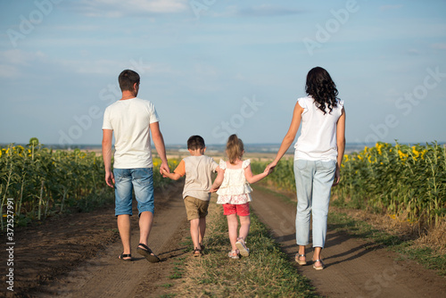 A family of four people mother, father, son and daughter walk in the countryside along a field with sunflowers. Family on a walk, view from the back. Family in summer clothes walking and holding hand