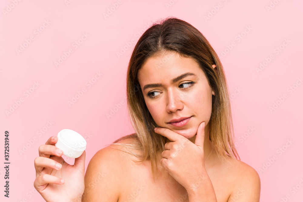 Young caucasian woman holding a moisturizer isolated looking sideways with doubtful and skeptical expression.