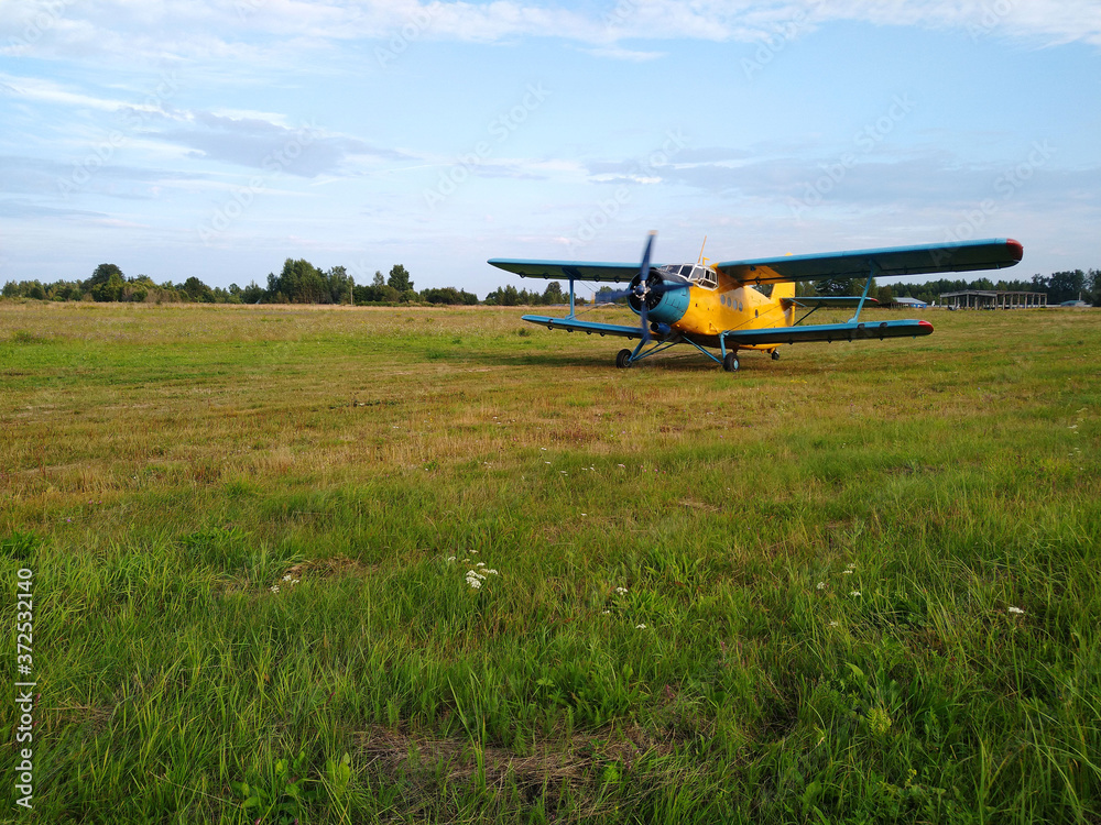 An old yellow biplane with blue wings and a running engine drives through a field of green grass in summer against a blue sky with white clouds An old yellow biplane with blue wings and a running engine drives through a field of green grass in summer against a blue sky with white clouds