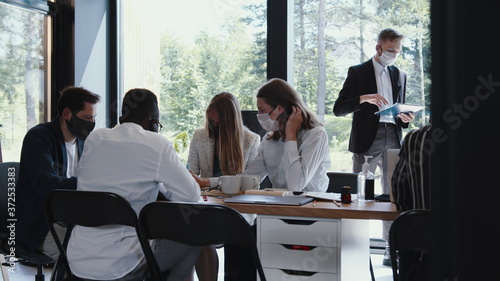 Team of diverse young happy business people working at modern office table wearing masks. Safety measures at workplace.