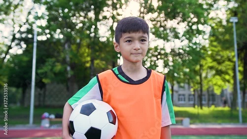 Handsome satisifed teen football player in orange vest standing with ball on the outdoors sport field before the beginning the training photo