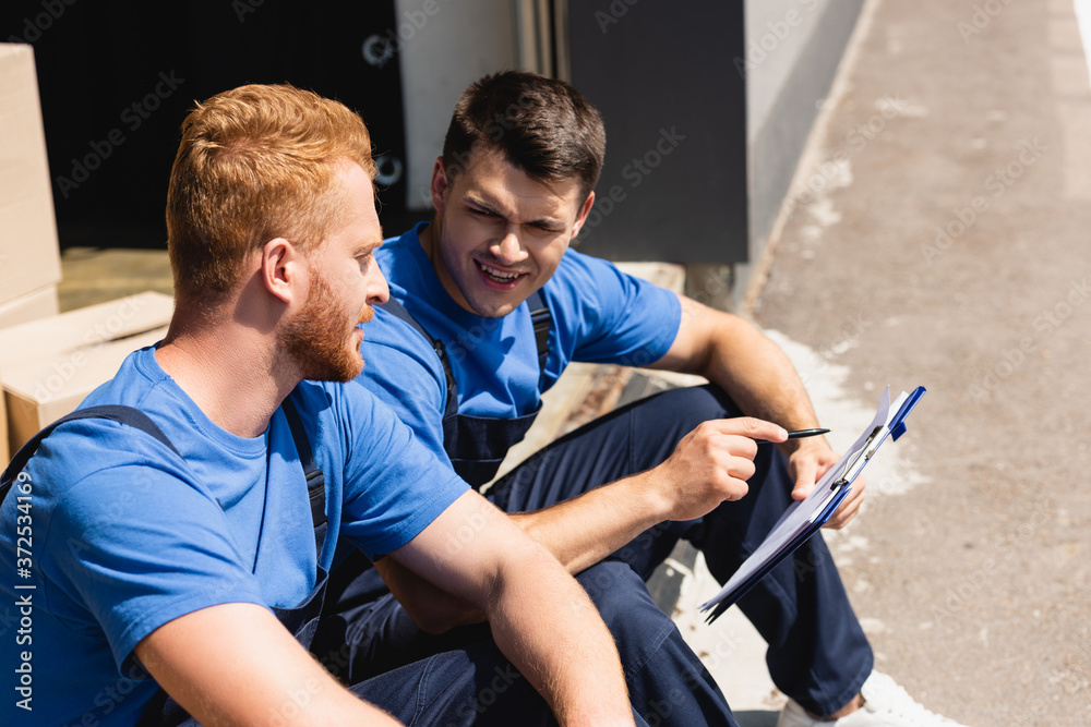 Obraz premium Loaders with pen and clipboard talking while sitting in warehouse outdoors