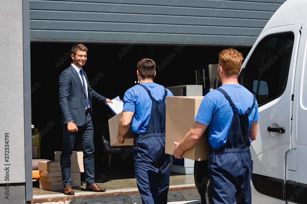 Selective focus of businessman holding clipboard while movers carrying packages near truck and warehouse outdoors