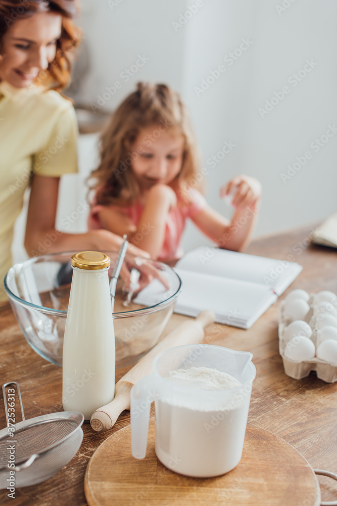 selective focus of mom and daughter reading cookbook near eggs, milk, flour, glass bowl and cooking utensils selective focus of mom and daughter reading cookbook near eggs, milk, flour, glass bowl and cooking utensils