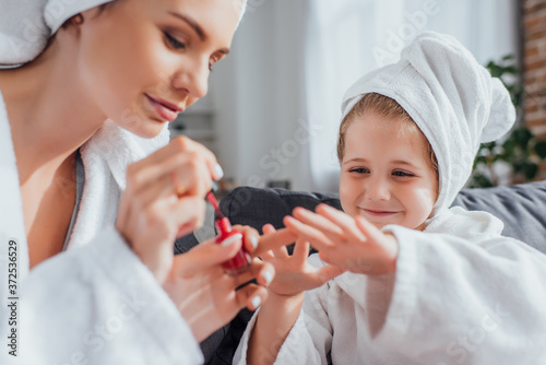 selective focus of young woman in white bathrobe covering fingernails of daughter with red enamel