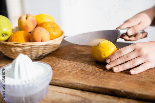 cropped view of woman cutting lemon on chopping board near fruits and juicer