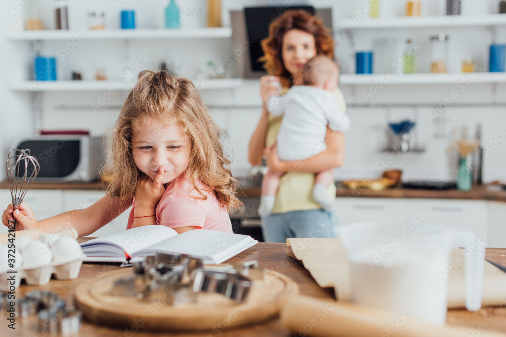 selective focus of girl holding whisk and reading cookbook near mother holding little son