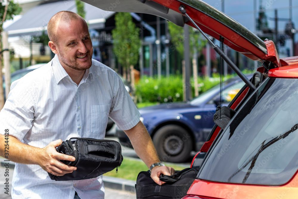 Photo of man putting bags in the car trunk. Businessman is unpacking the car trunk.Man packing luggage in car ready for road trip.Young Caucasian man loading the trunk of the car parked on the parking