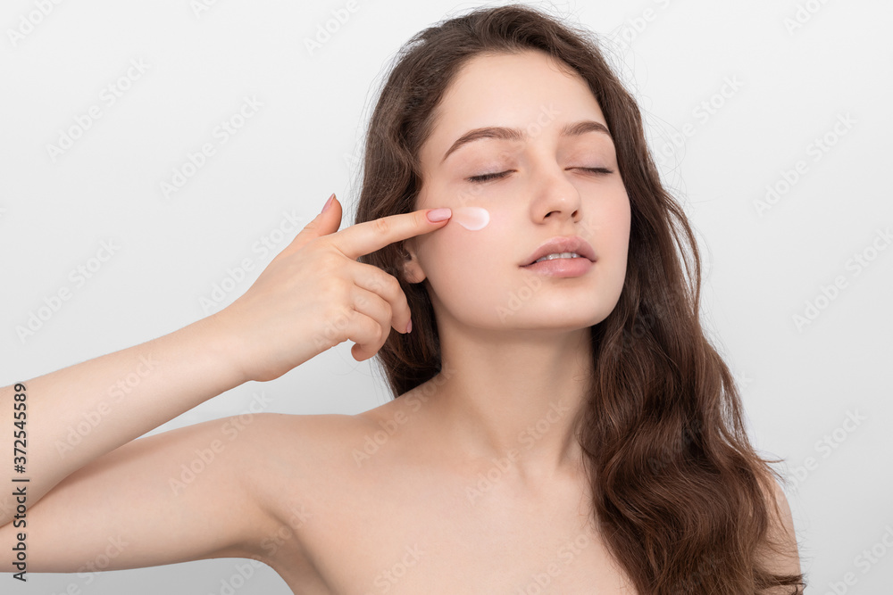 Portrait of young woman applying moisturizer cream on her face. Young girl with long hair and blue eyes. Portrait of young woman applying moisturizer cream on her face. Young girl with long hair and blue eyes.