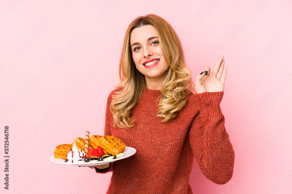 Young blonde woman eating a waffle dessert isolated cheerful and confident showing ok gesture. Young blonde woman eating a waffle dessert isolated cheerful and confident showing ok gesture.