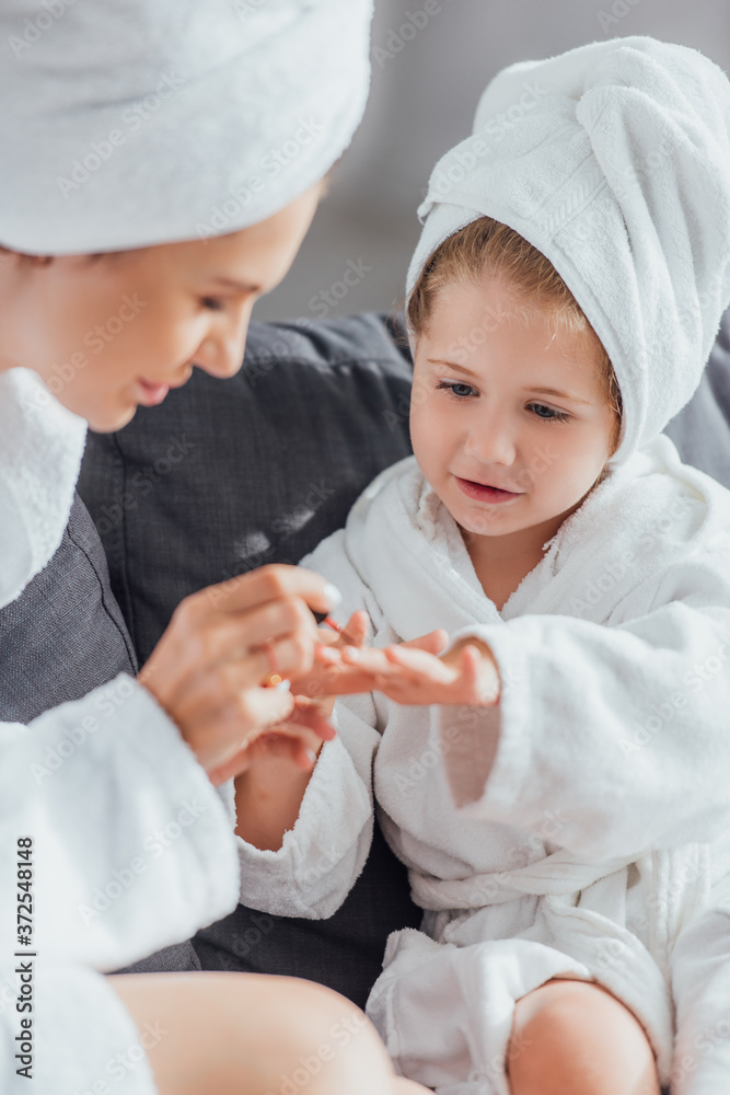 selective focus of mother applying enamel on fingernails of daughter while sitting together in white bathrobes