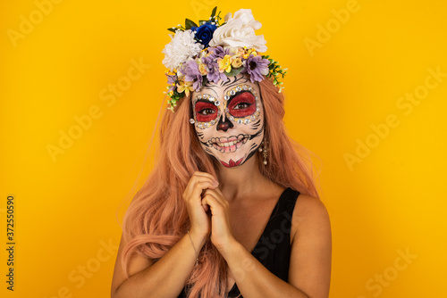 Dreamy charming young female sugar skull make up with pleasant expression, keeps hands crossed near face, excited about something pleasant.