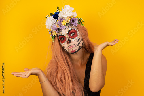 Beautiful woman wearing halloween make up doubt expression, confuse and wonder concept, uncertain future shrugging shoulders, standing against gray Wall.