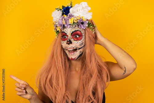 Portrait of young woman wearing halloween make up with shocked facial expression, showing something amazing on blank space, one hand on her head and pointing with forefinger. OMG concept.