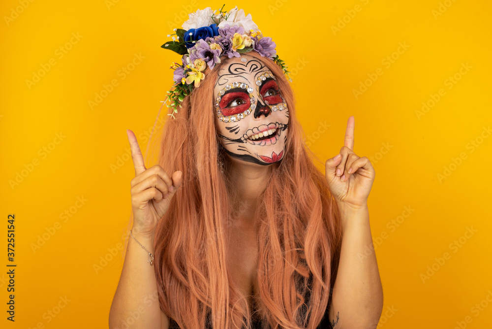 Horizontal shot of successful friendly looking young dark haired woman with bright make up exclaiming excitedly, pointing both index fingers up, indicating something shocking on blank copy space wall. Horizontal shot of successful friendly looking young dark haired woman with bright make up exclaiming excitedly, pointing both index fingers up, indicating something shocking on blank copy space wall.