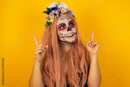 Horizontal shot of successful friendly looking young dark haired woman with bright make up exclaiming excitedly, pointing both index fingers up, indicating something shocking on blank copy space wall.