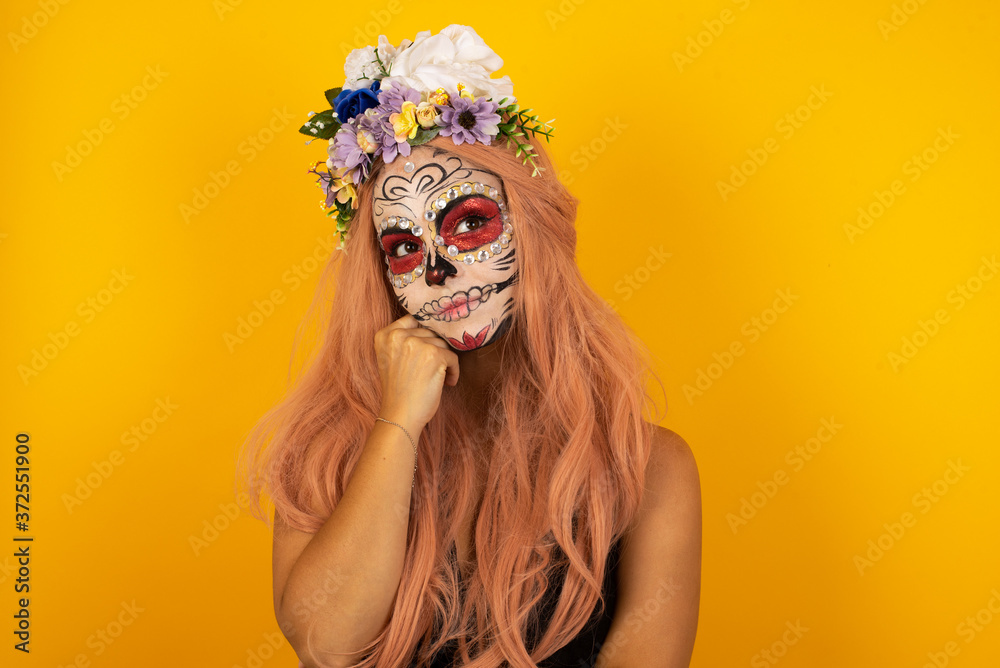 Face expressions and emotions. Thoughtful young pretty girl wearing sugar skull Halloween make up holding hand under her head, having doubtful look.