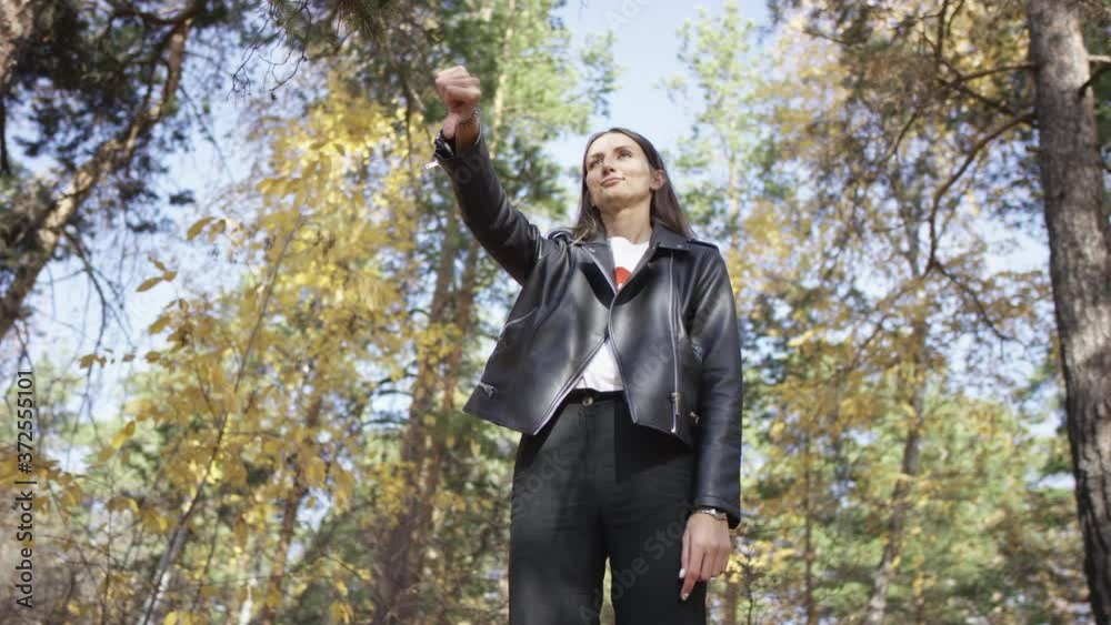 4K portrait of sceptical long-haired brunette grimacing disapprovingly putting out tongue and giving thumbs up standing in autumn forest. Low angle 360 degree tracking arc shot.