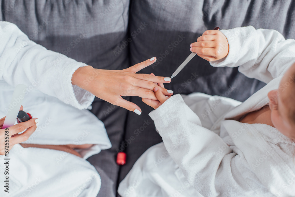top view of child making manicure with nail file to mother while sitting on sofa in white bathrobes together top view of child making manicure with nail file to mother while sitting on sofa in white bathrobes together