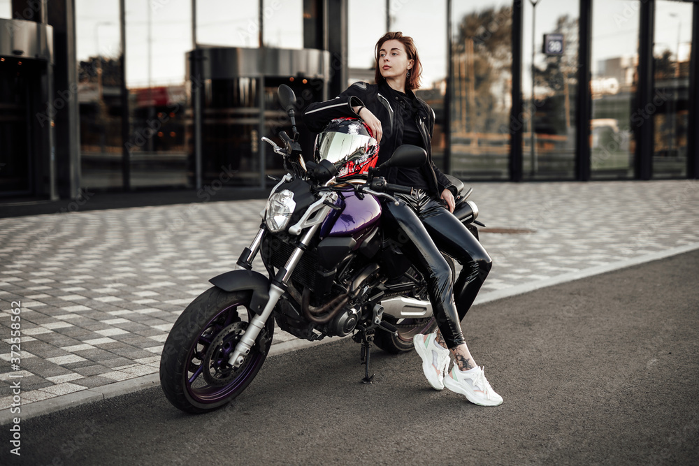 Stylish girl in a black jacket sits on a purple motorbike with a red safety helmet